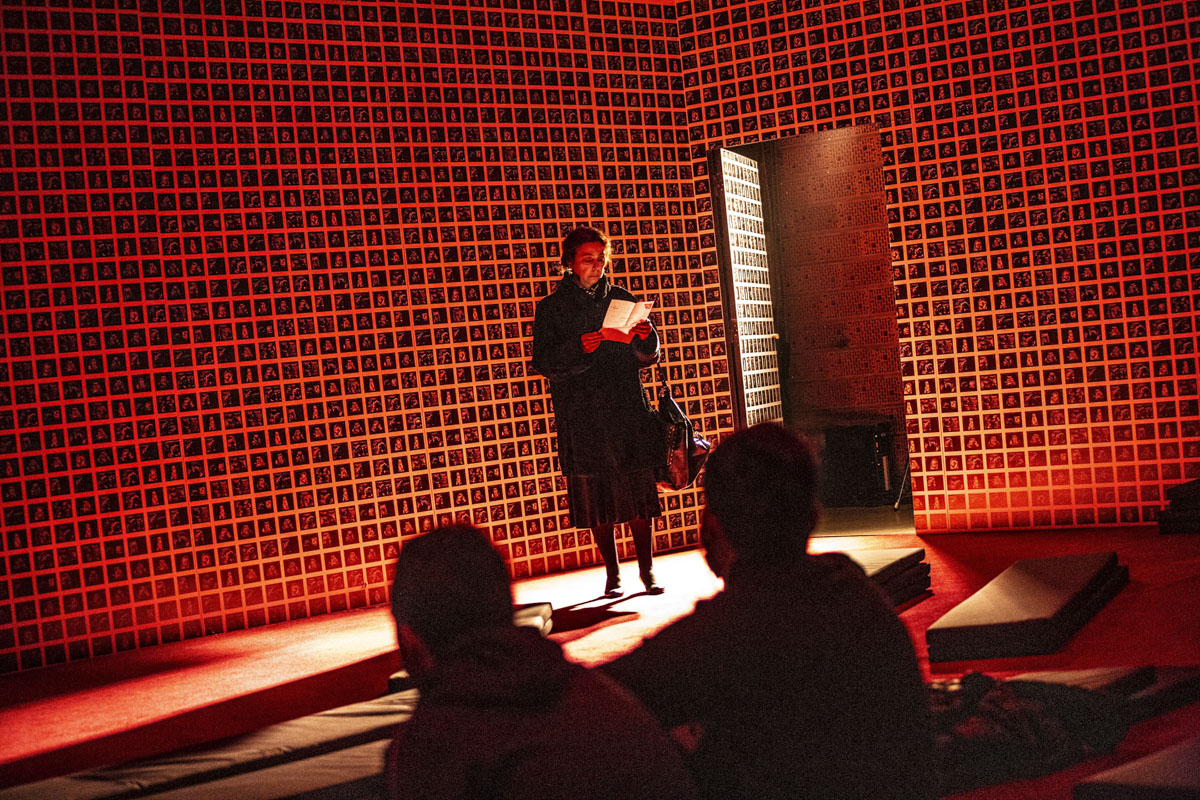 Starsza kobieta w czarnym futerku stoi na tle podświetlonej terakotowej ściany. Z uwagą czyta pismo z zawiadomieniem.