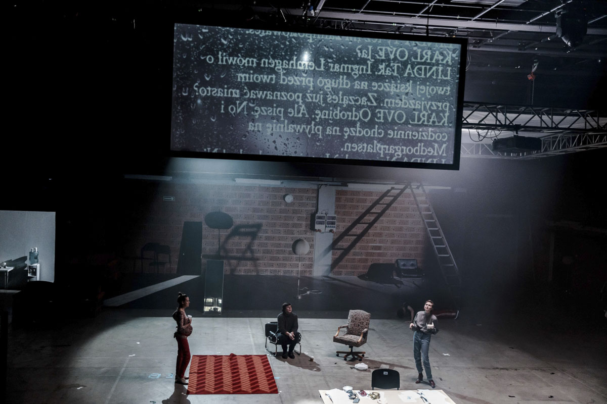 Na scenie stoją dwie kobiety. Między nimi na fotelu siedzi młody mężczyzna. Pod sufitem zawieszony ekran gdzie wyświetlany jest fragmentem tekstu sztuki.