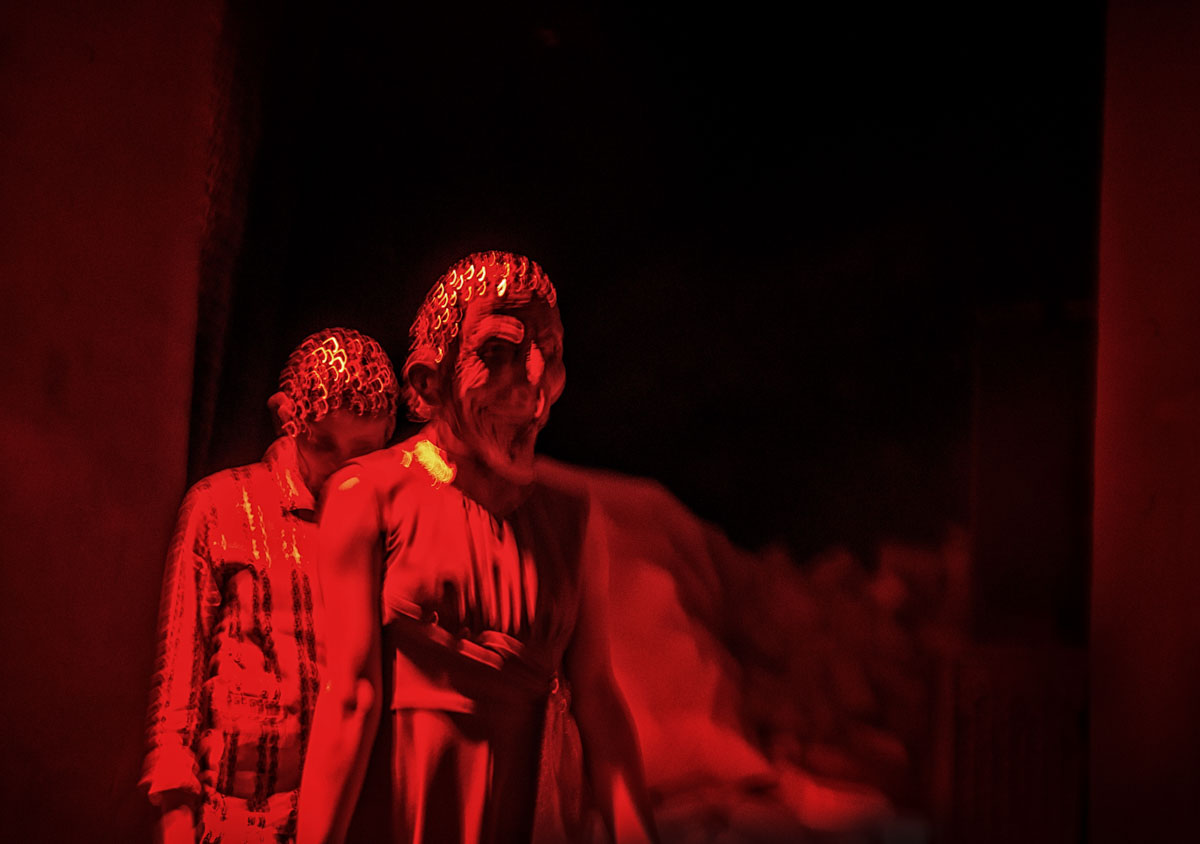 W czerwonym świetle dwie osoby od pasa w górę, jedna za drugą. Na twarzy pierwszej maska z szeroko otwartymi ustami.
