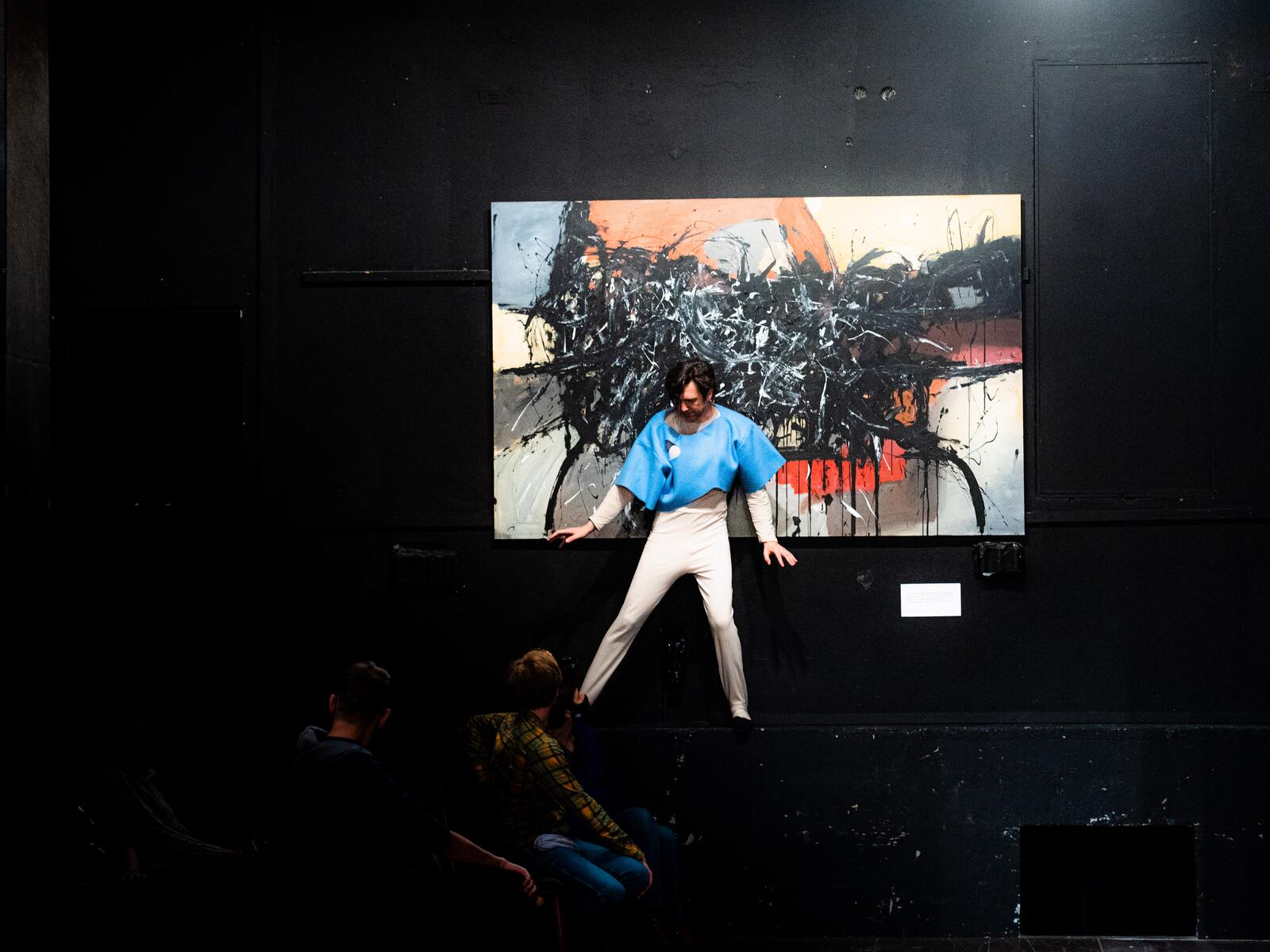 Na występie muru stoi w rozkroku mężczyzna w beżowym trykocie i niebieskiej krótkiej tunice. Za nim abstrakcyjny obraz.