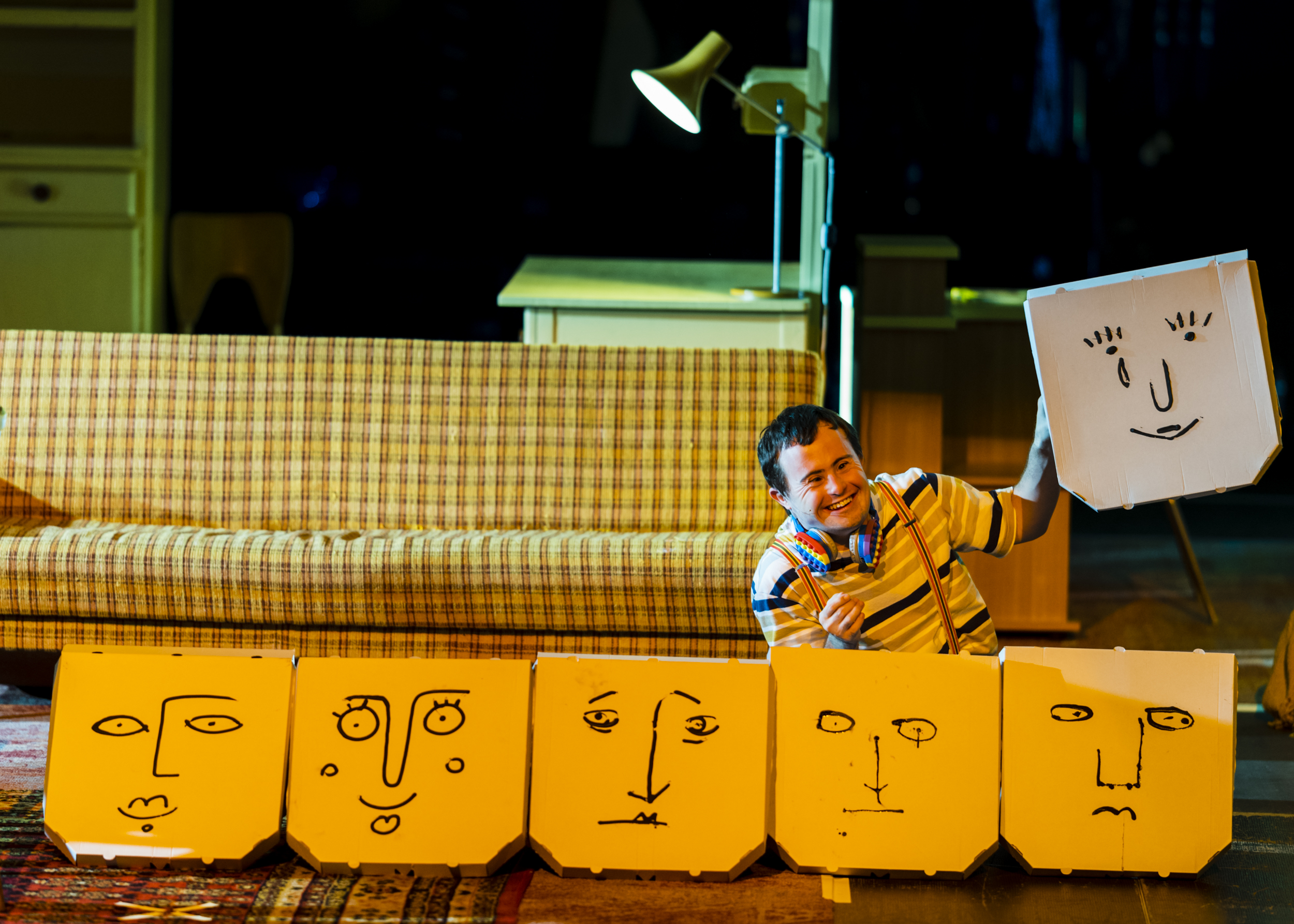 W rzędzie na podłodze ustawione są pudełka do pizzy z narysowanymi dziecięcą kreską twarzami wyrażającymi różne emocje. Za pudełkami siedzi rozbawiony mężczyzna, który w uniesionej dłoni trzyma pudełko do pizzy z narysowaną uśmiechniętą twarzą i lecącą z oka łezką.